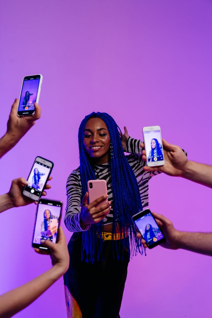 gallery-5 Portrait of a stylish woman with blue hair taking selfies surrounded by smartphones in a studio setting.